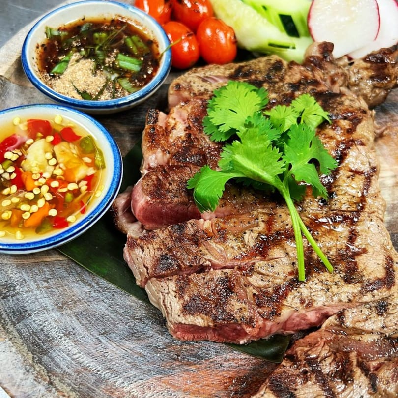 Grilled Thai steak with two dipping sauces, cherry tomatoes, cucumber, and radish on a wooden board.
