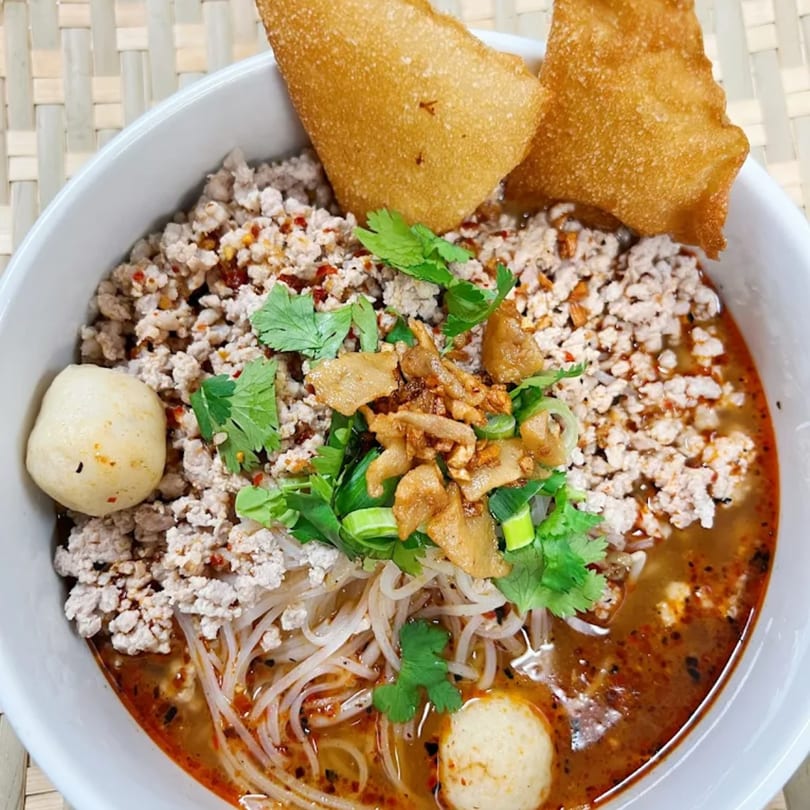 A bowl of Thai Tom Yum noodle soup with minced pork, fish balls, and crispy wontons.
