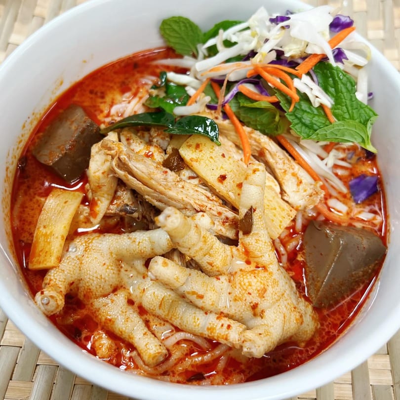 Spicy red curry noodle soup with chicken feet, shredded chicken, and fresh herbs in a white bowl.